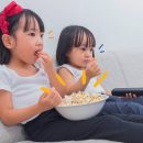 Foto de duas meninas sentadas no sofá assistindo a um filme. A maior delas segura uma bacia de pipoca e a outra segura o controle remoto