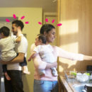 Um homem e uma mulher cada com um bebê no colo em uma cozinha. A mulher mexa na pia e homem na geladeira.