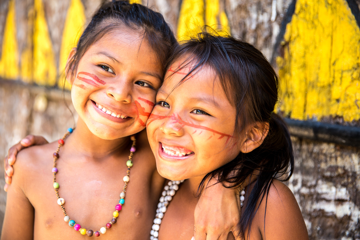 Duas meninas indígenas abraçadas e sorrindo, com o rosto pintado e ornamentos típicos da Amazônia pendurados no pescoço.