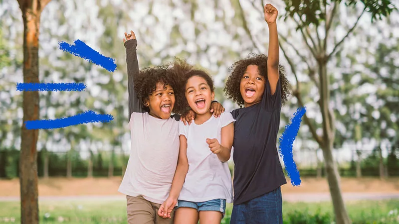 Três crianças abraçadas. sorrindo, e com as mãos levantadas para o alto, em sinal de comemoração e alegria.