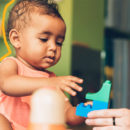 Criança pequena com roupa rosa brinca com um brinquedo de montar. Uma mãe de adulto aparece segurando o brinquedo