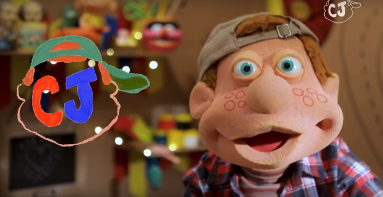 Personagem Júlio, do programa de TV Cocoricó, aparece em um cenário com fundo desfocado, com brinquedos e objetos coloridos.