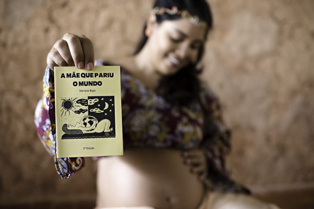 A poeta Mariane Bigio grávida segurando o cordel A mãe que pariu o mundo com uma das mãos, e com a outra alisando a barriga.