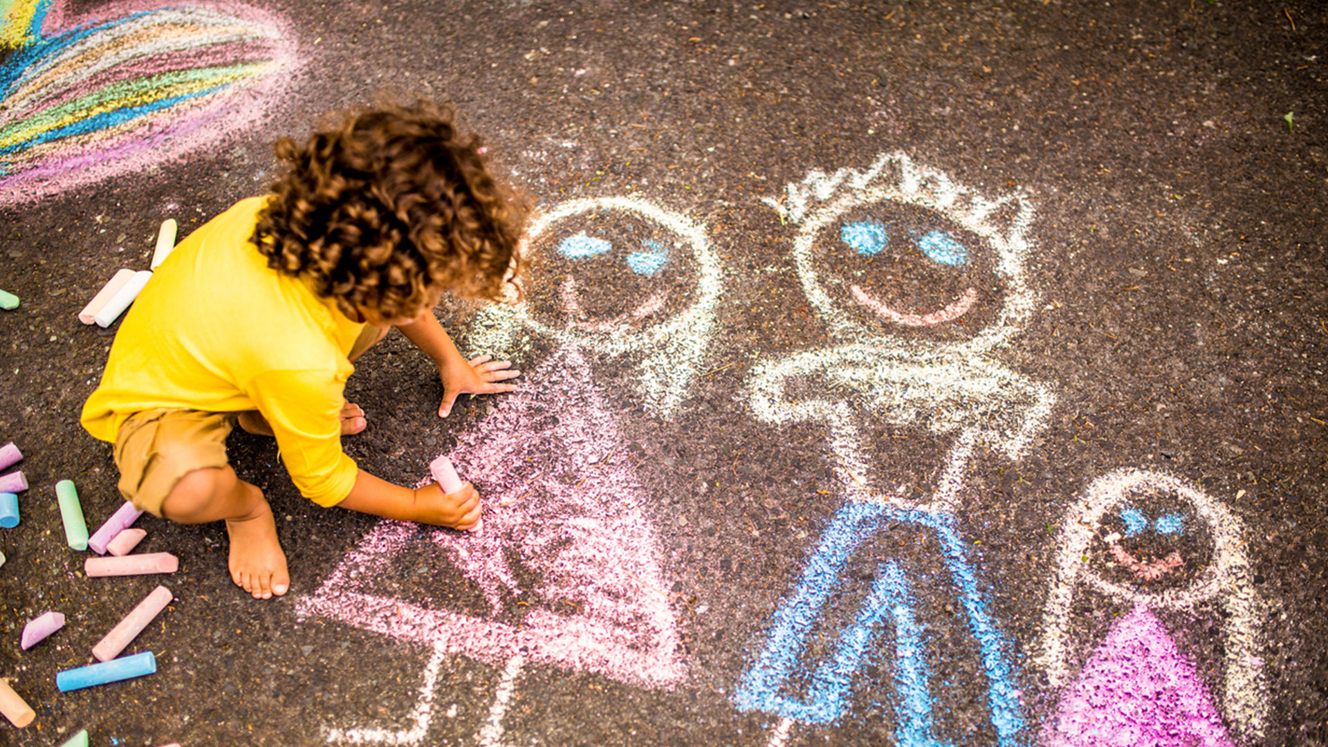 Menino desenhando uma ciranda de bonequinhos coloridos com giz de lousa no asfalto da rua