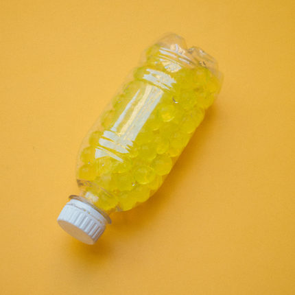 Em um fundo amarelo vê-se uma garrafa pet transparente cheia de bolinhas amarelas