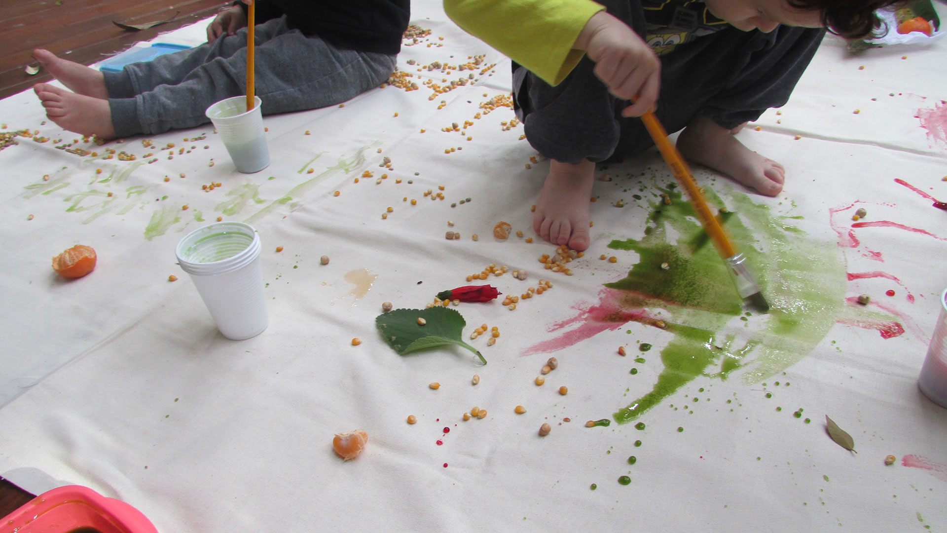 Crianças pintando um painel branco estendido no chão com tintas coloridas e pincéis com aquarelados.