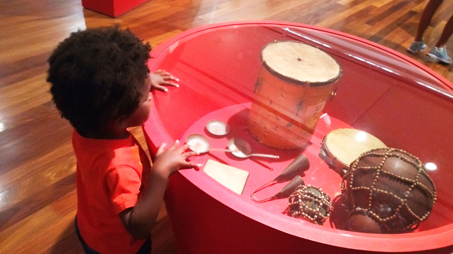 Menino negro explorando objetos de exposição afro-brasileira: instrumentos musicais como agogô, pandeiro e xequerê dispostos em uma mesa vermelha com tampo de vidro