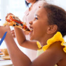 Menina comendo pedaço pizza com as mãos, sorrindo e olhando para frente.