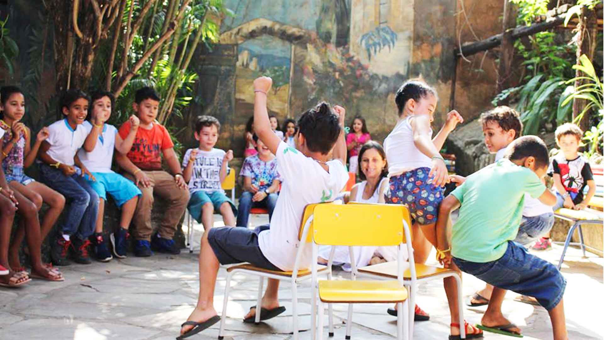 Foto de um grupo de mais de 10 crianças, em círculo, brincando felizes de dança das cadeiras