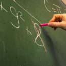 Imagem de uma mão escrevendo em uma lousa. Na lousa contém uma expressão matemática, e o número 8 da expressão está com um X