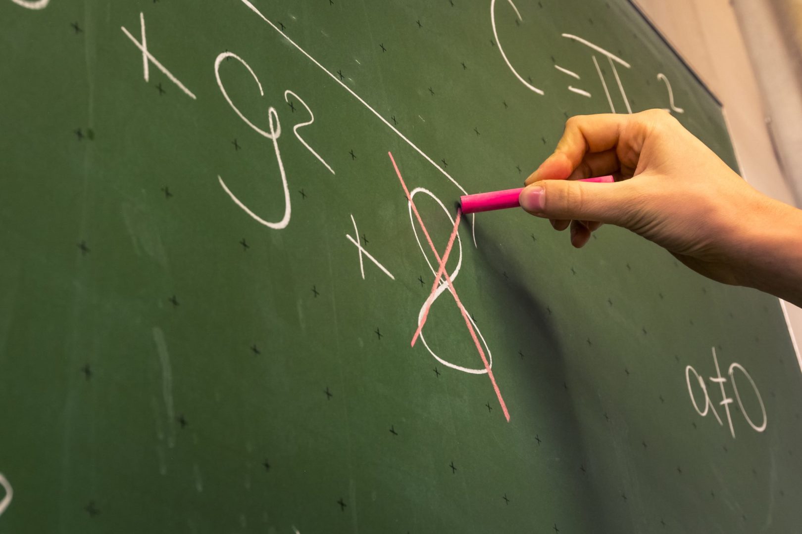 Imagem de uma mão escrevendo em uma lousa. Na lousa contém uma expressão matemática, e o número 8 da expressão está com um X