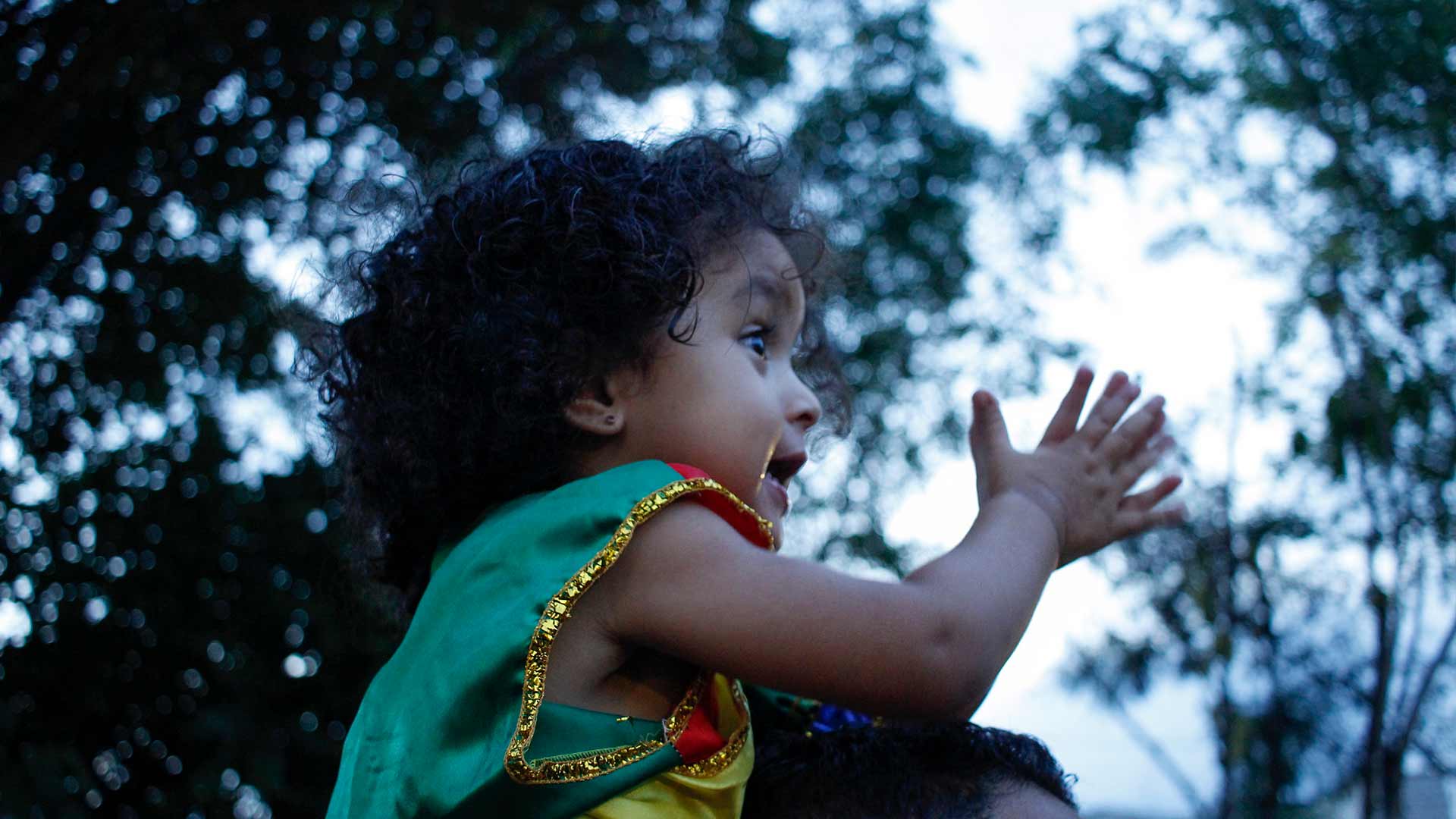 Foto de uma criança batendo palmas, feliz, com roupa de carnaval no ombro de um adulto