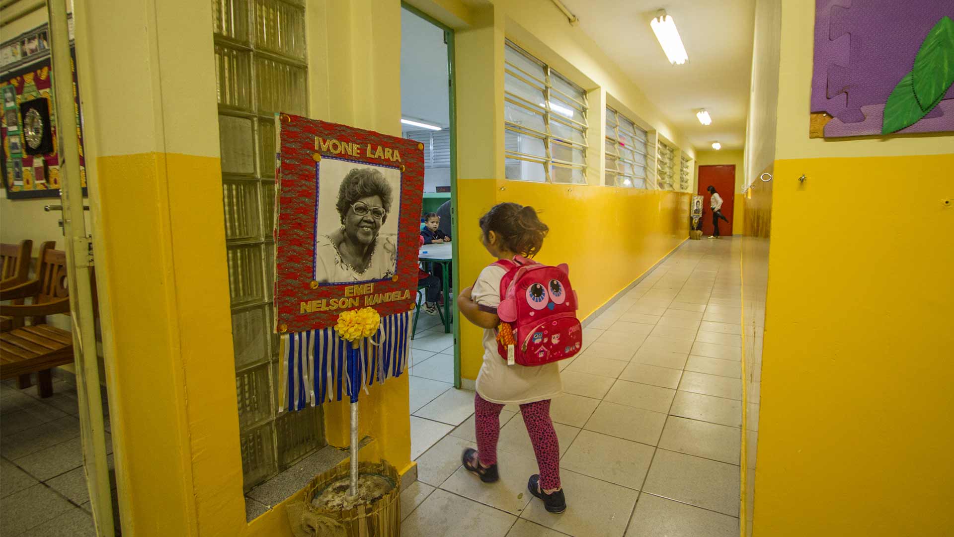 Foto da entrada da sala de aula, tem uma menina entrando em sala de aula, com sua mochila rosa, e ao lado da entrada da sala, uma placa com o retrato da Dona Ivone Lara