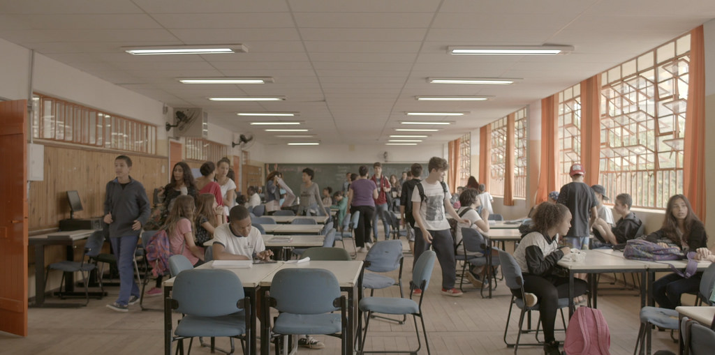Foto em uma sala de aula, alguns alunos sentados, outros em pé