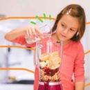 Imagem de uma menina colocando água no liquidificador que contém frutas picadas