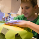 Foto de um menino com uma máquina de costura, a mão dele está em cima do pano verde fazendo a costura