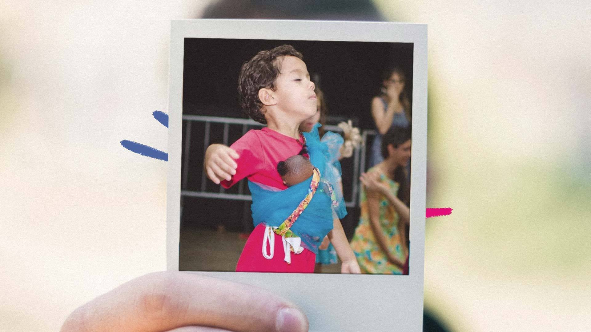 Imagem de um menino com os olhos fechados e com a mão suspensa dançando levemente, e carregando uma boneca negra amarrada em seu corpo.