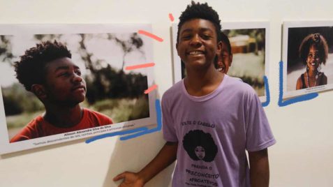 Foto de um menino negro sorrindo com uma camiseta escrita “solte o cabelo”, ele está ao seu lado tem um retrato colado na parede de uma criança negra.