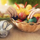 Foto de uma cesta de ovos pintados e duas mãos pegando os ovos. A cesta está sobre uma mesa juntamente com um coelho