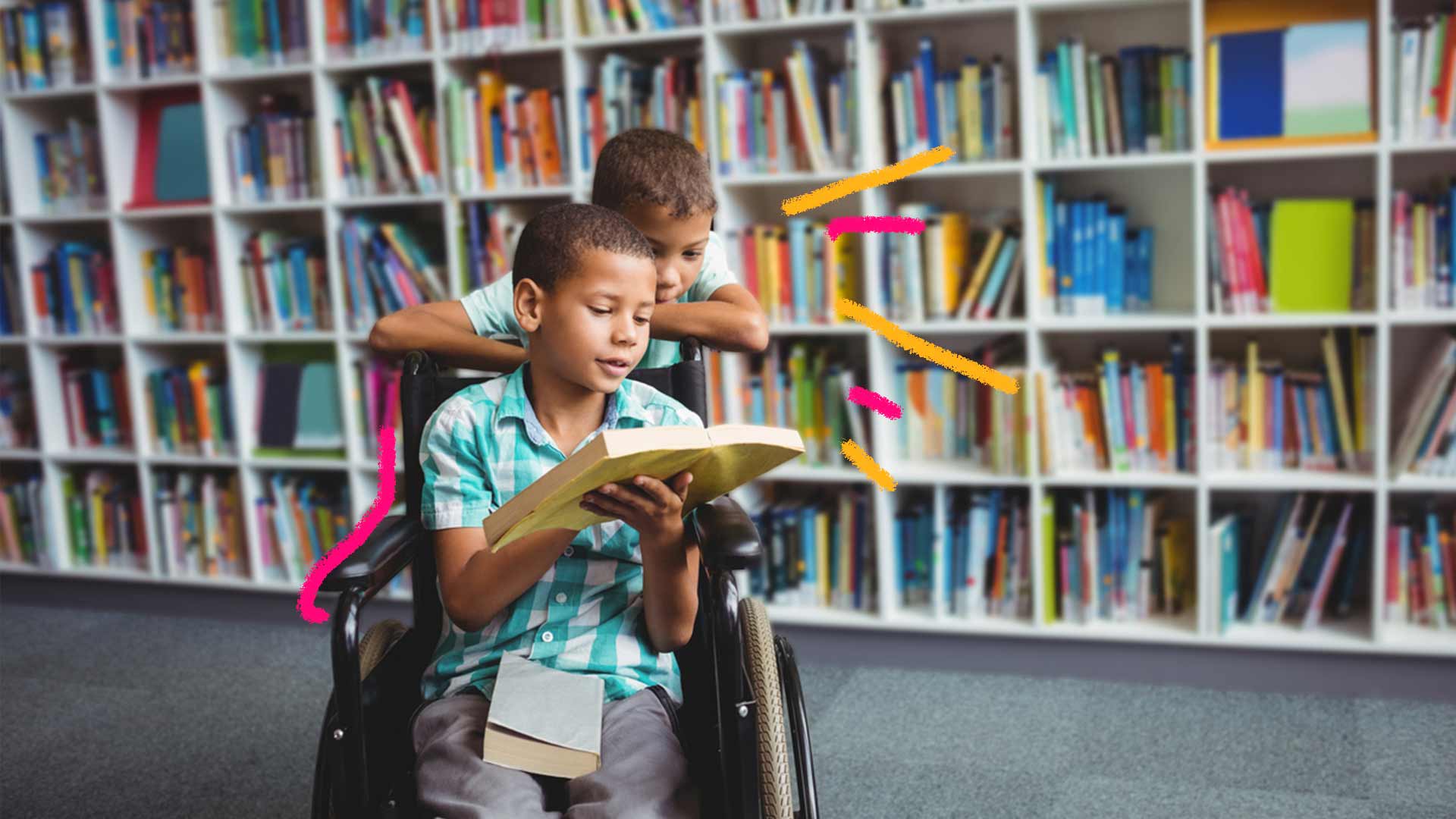Foto de um menino na cadeira de rodas com um livro em mãos e com ele, outro menino apoiado na cadeira de rodas, ambos estão em uma biblioteca.