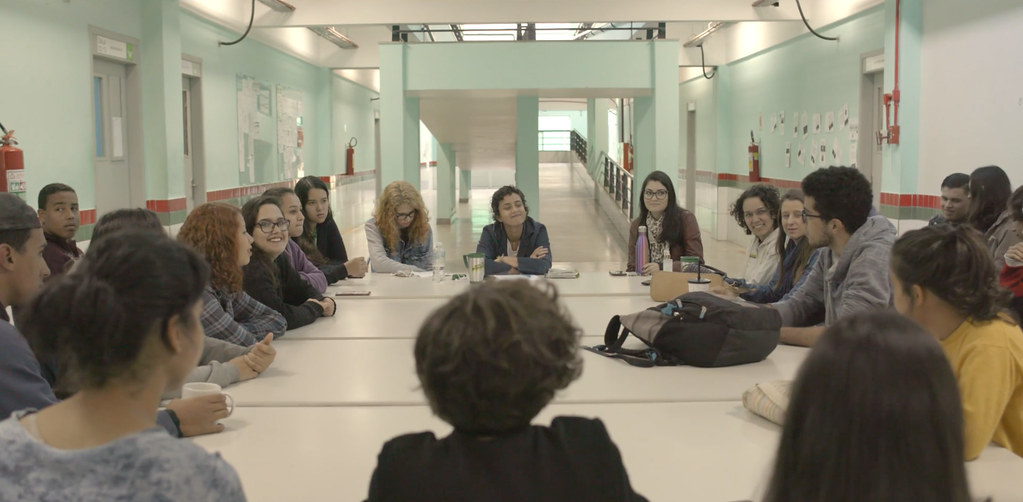 Foto de várias professoras sentandas em uma mesa de reunião, conversando. Elas estão dentro da escola