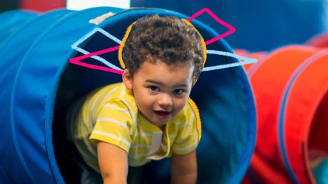 Foto de um menino engatinhando e brincando saindo de um tubo azul com a expressão feliz