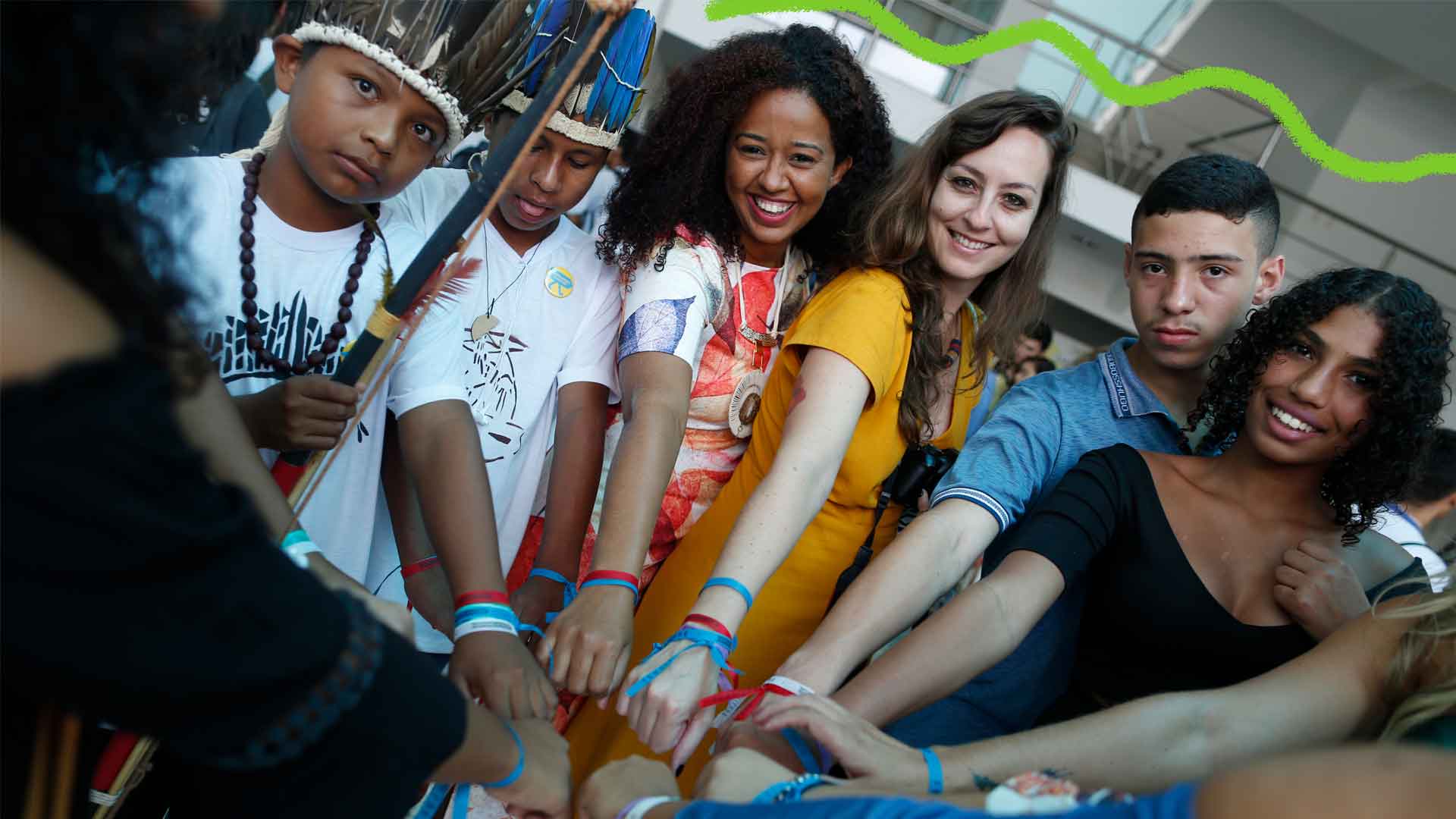 Foto de vários jovens juntas sorrindo e unindo seus braços para participarem do projeto Criativos da Escola.