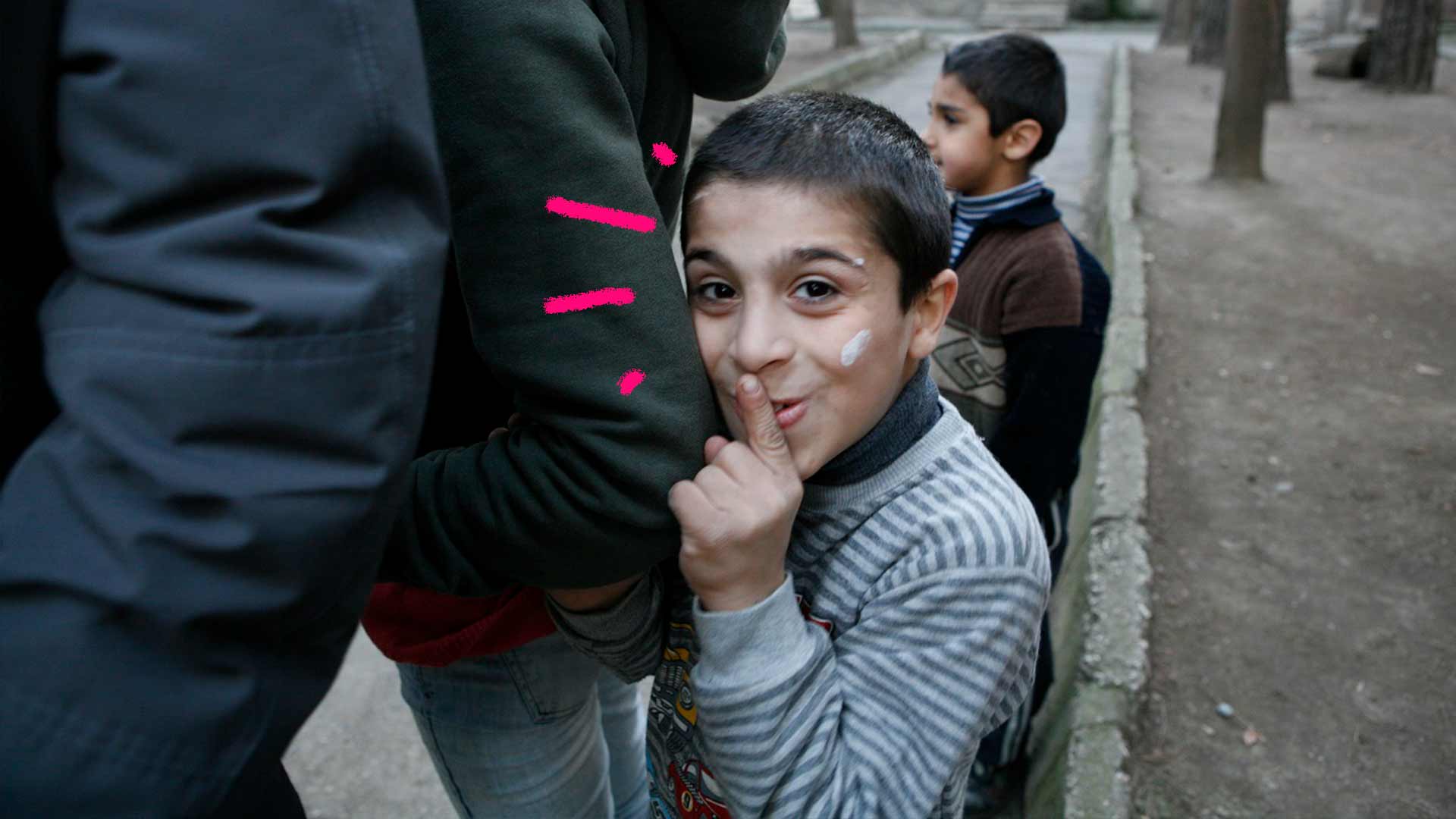Foto de um menino com o dedo na boca fazendo sinal de silêncio. O menino está escondido atrás de um adulto com os braços dados