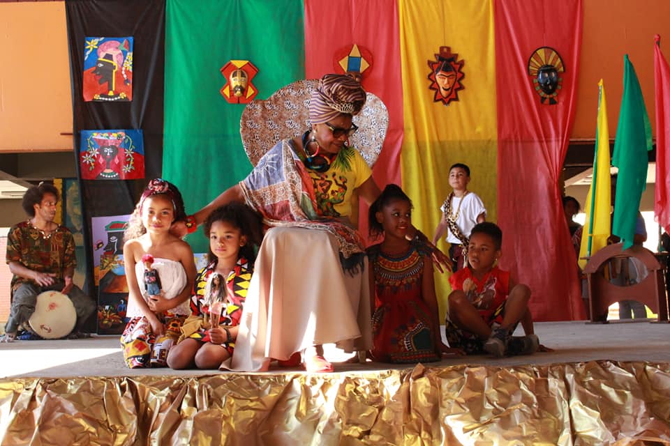 Foto de quatro crianças com uma professora Quilombela. Todos estão sentados em um palco colorido e a professora está em sua cadeira olhando e tocando nas crianças.