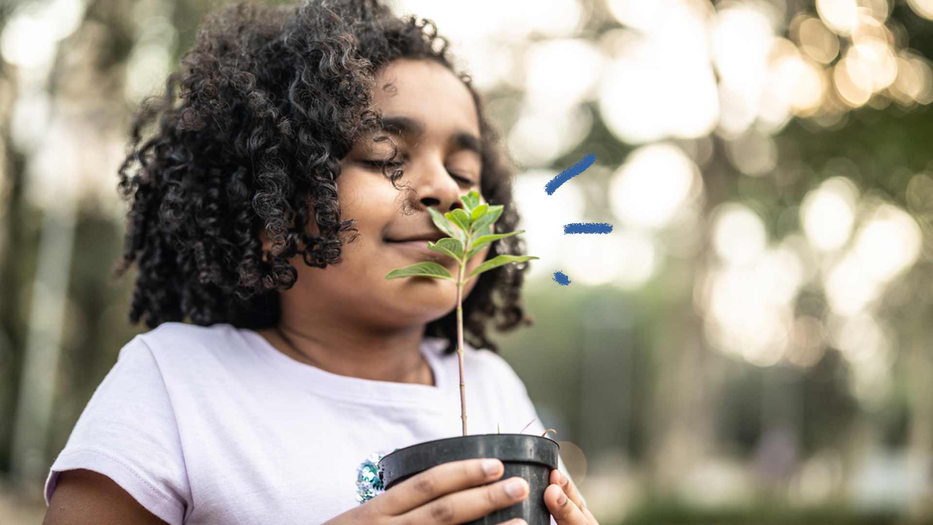 Foto de uma criança negra com os olhos fechados cheirando um vaso de planta em suas mãos