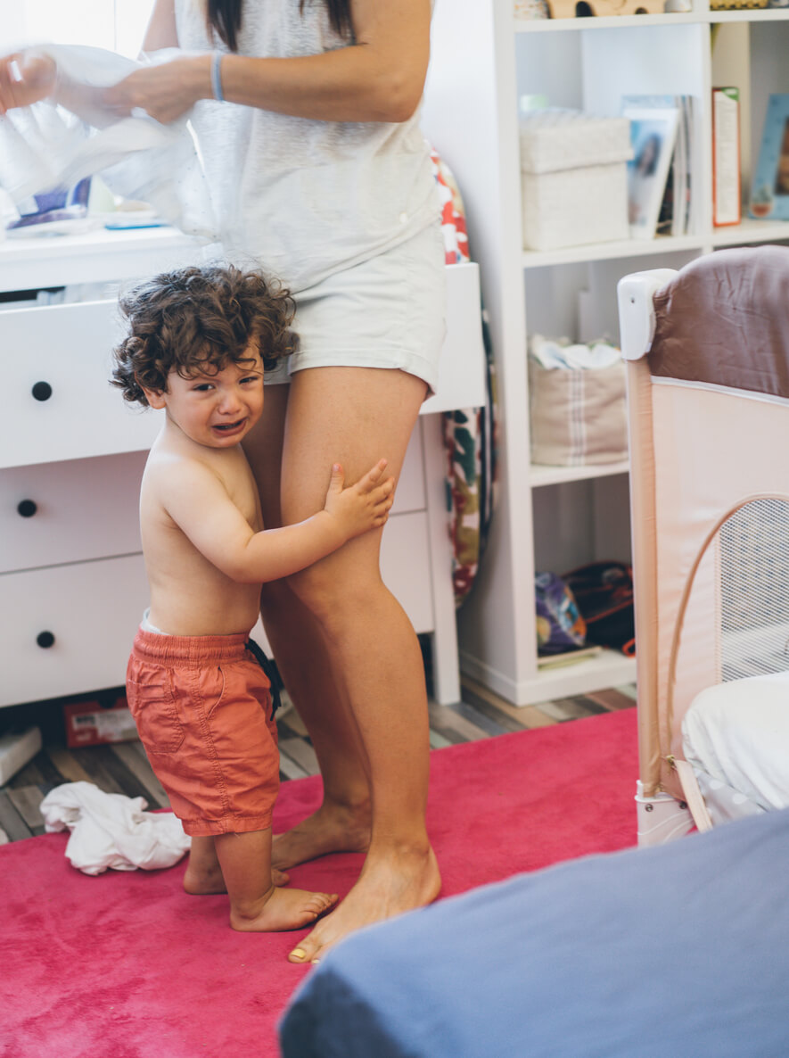 Foto de um quarto bagunçado, uma criança de aproximadamente dois anos agarra as pernas de uma mulher, cuja imagem só aparece da cintura para baixo. A criança chora e olha para a câmera, enquanto a mulher tenta se esquivar.