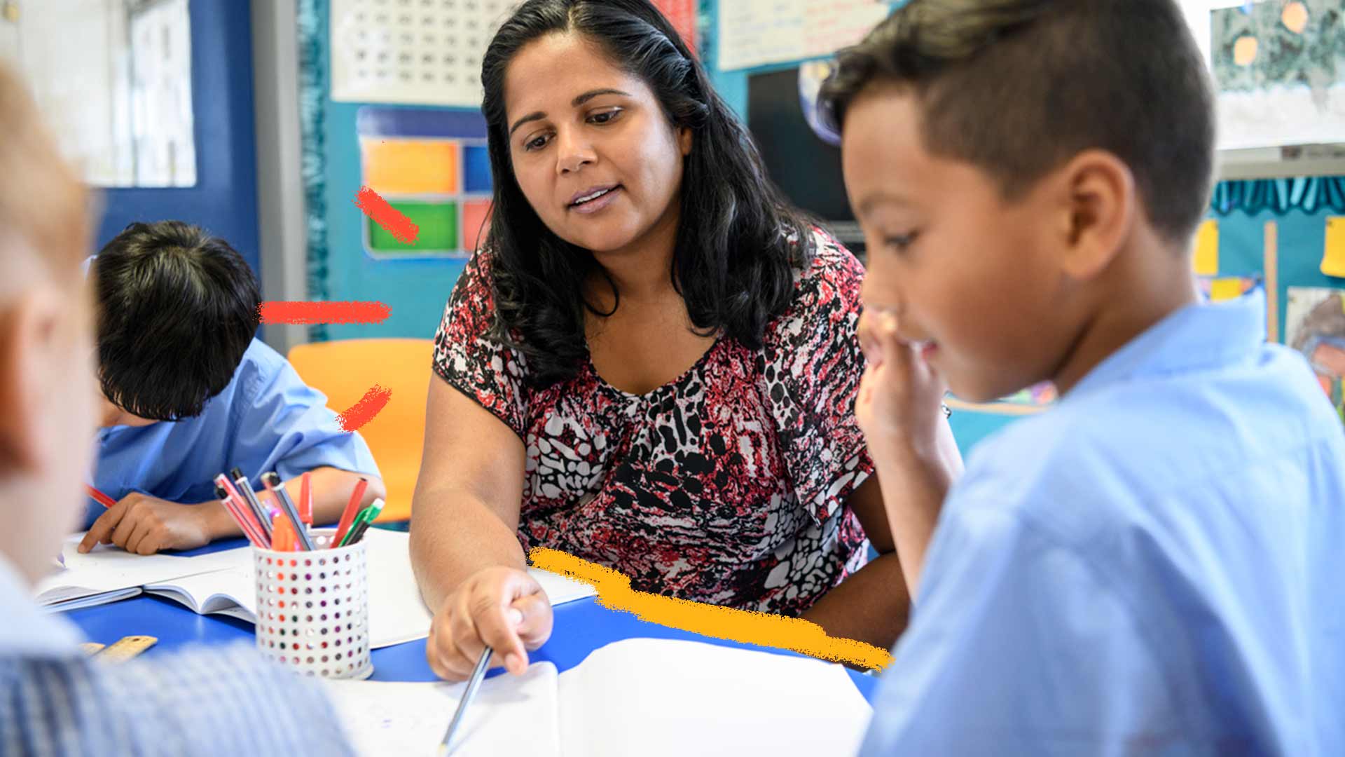 Foto de uma professora com três alunos sentados em sala de aula. Ela aponta para o caderno do menino