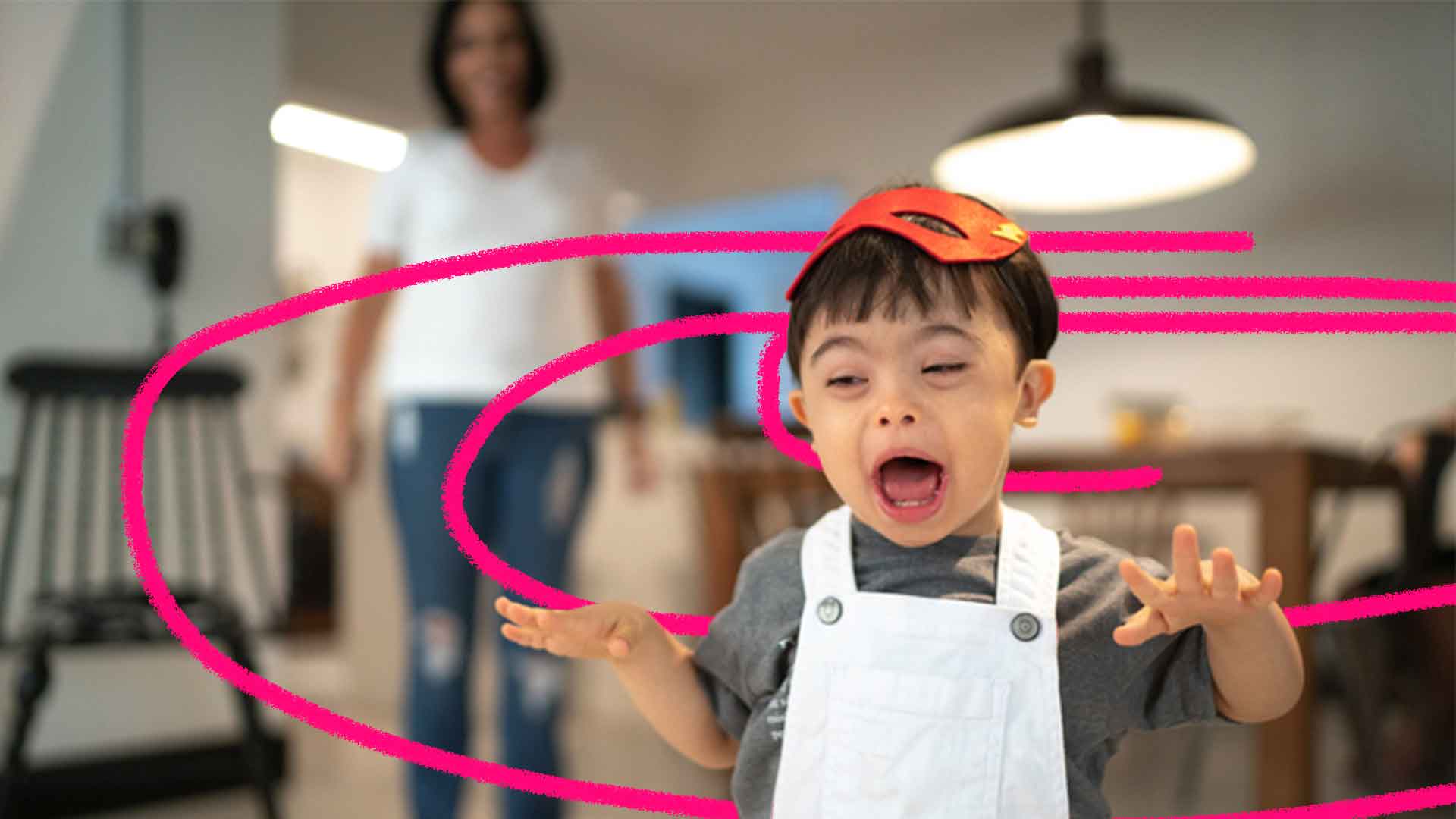 Imagem de uma criança em primeiro plano com síndrome de Down com a boca e as mãos levantadas. Em segundo plano, há uma mulher observando-o