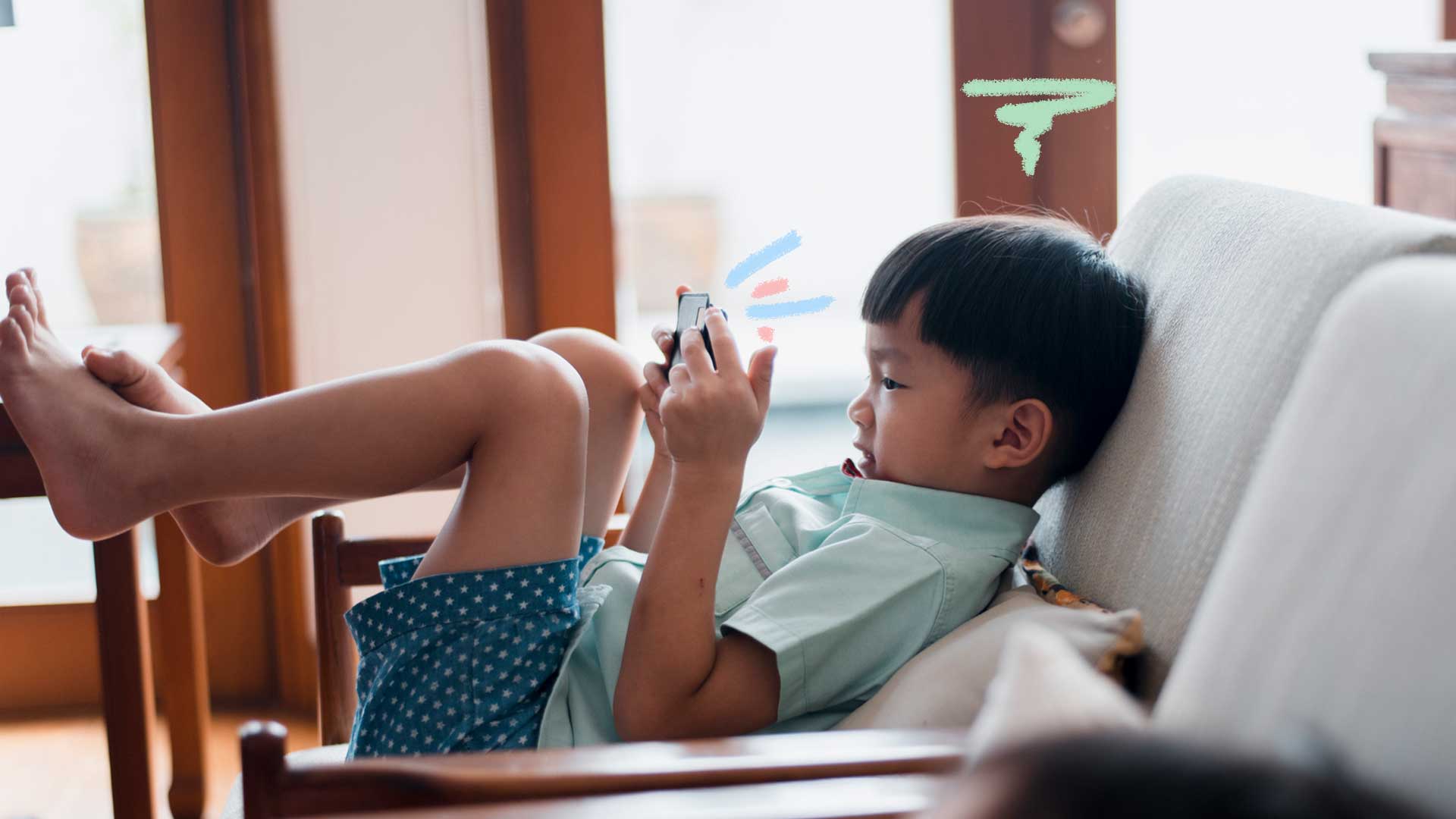 Imagem de um garoto sentando no sofá com as pernas para cima. Ele esta com um celular em mãos jogando.