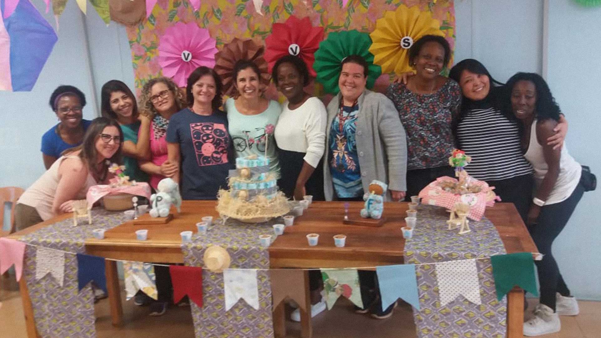 Foto de onze mulheres em pé em frente a uma mesa decorada com bolo, docinhos, lembrancinhas. Elas estão comemorando um chá de vó.