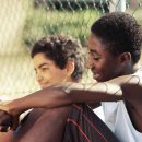 Imagem de dois garotos negros sentados e encostado conversando em uma grade de rede