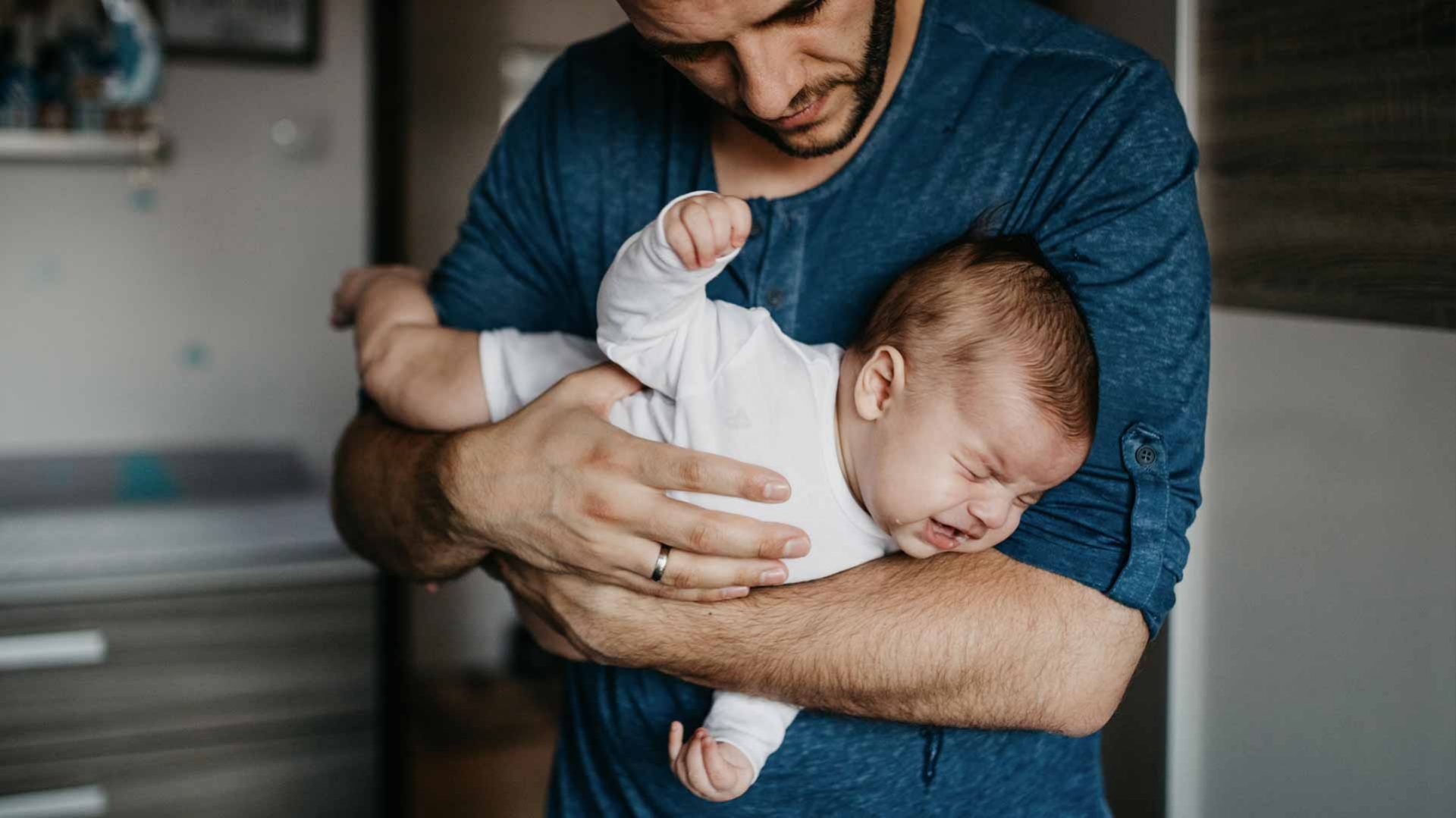 Imagem de um homem segurando o bebê em seus braços, o bebê esta de bruço e chorando.