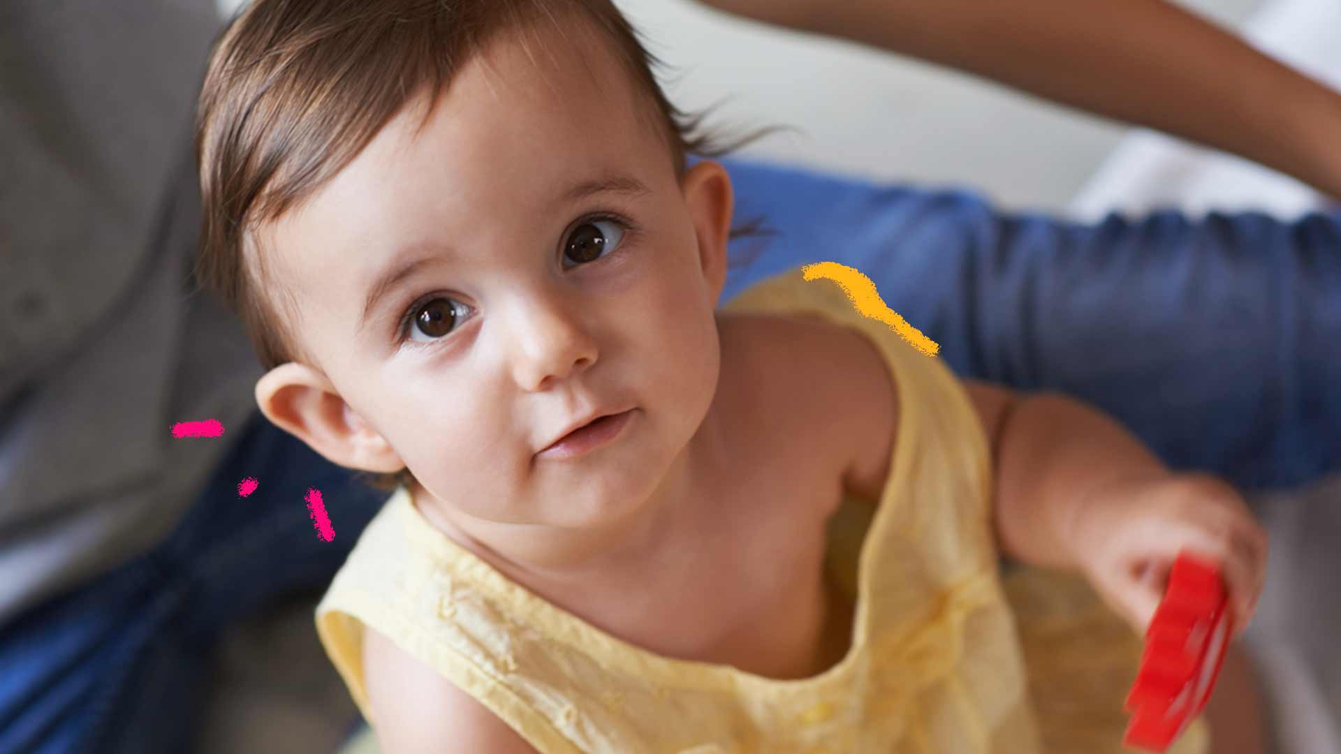 Foto de um bebê de body amarelo. Ele está sentado no chão e olha para a câmera que está posicionada acima de sua cabeça.
