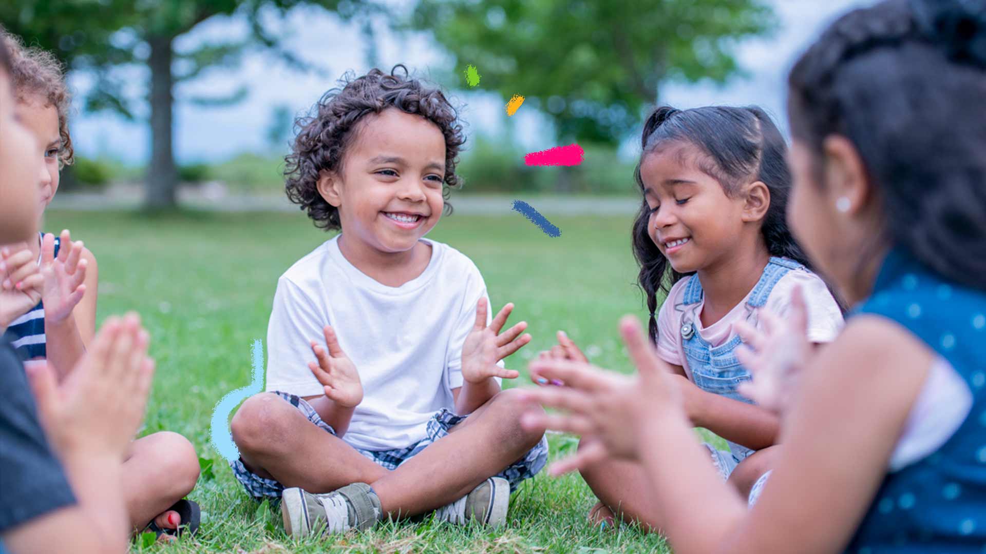 Imagem de cinco crianças sentadas em fila em um gramado, brincando de bater palmas. No centro da foto, um menino em destaque sorri e abana as mãos.