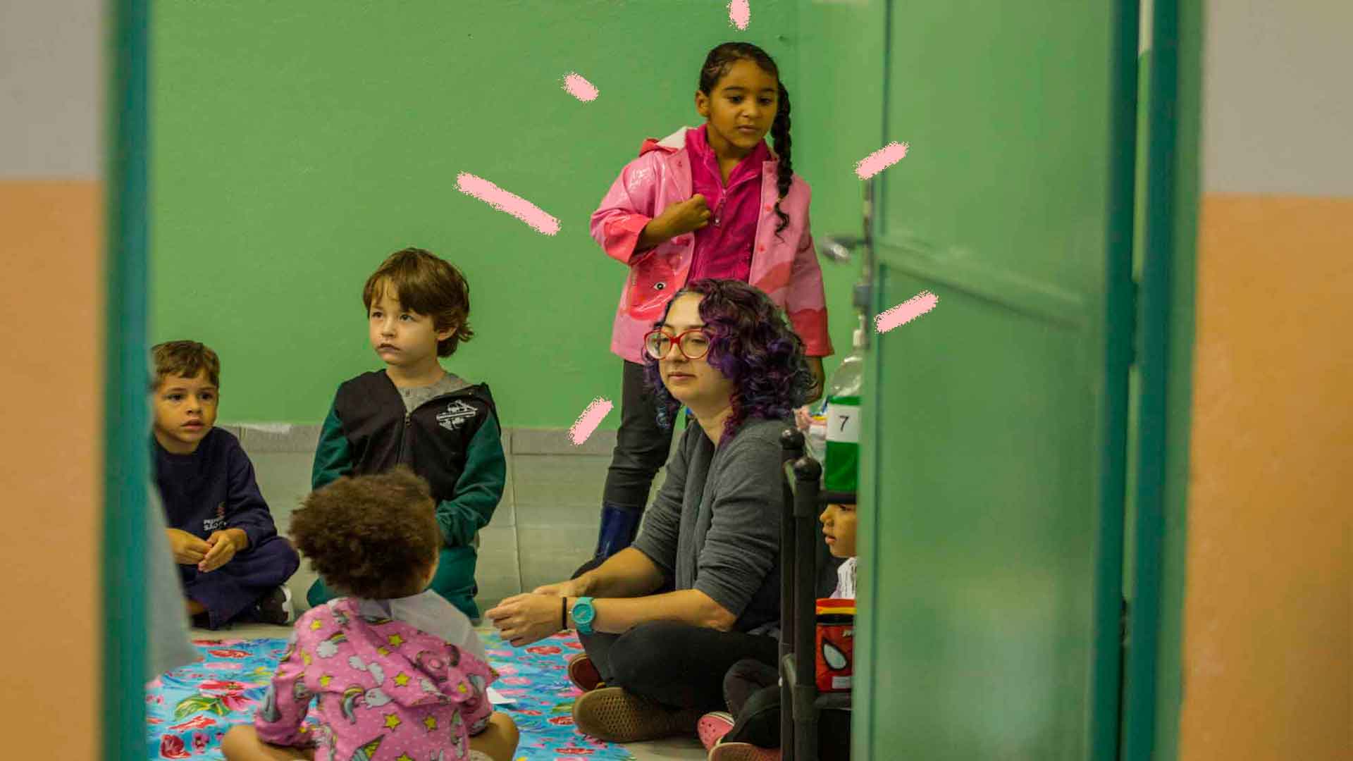 Foto de crianças com a professora sentados no chão. Em uma sala de aula com a cor da parede verde, elas observam atentamente a professora.