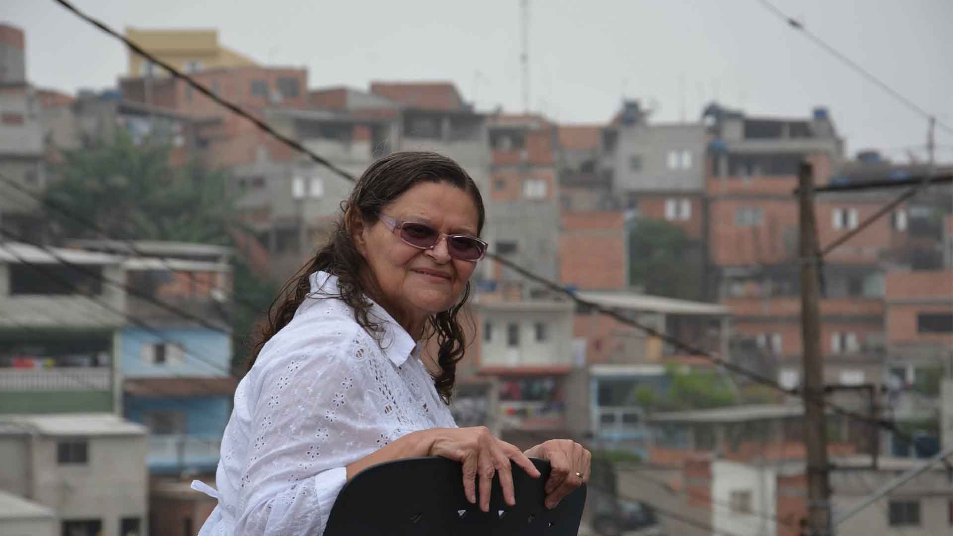 Uma mulher de óculos olha para a câmera. Ao fundo vê-se a favela