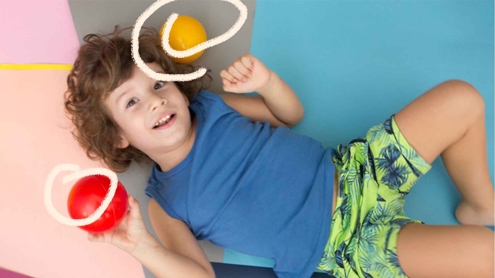 Foto de um menino deitado olhando para cima com uma bola vermelha na mão e uma amarela próxima da cabeça. Ela tem cabelo cacheado e veste uma camiseta azul e um short verde.