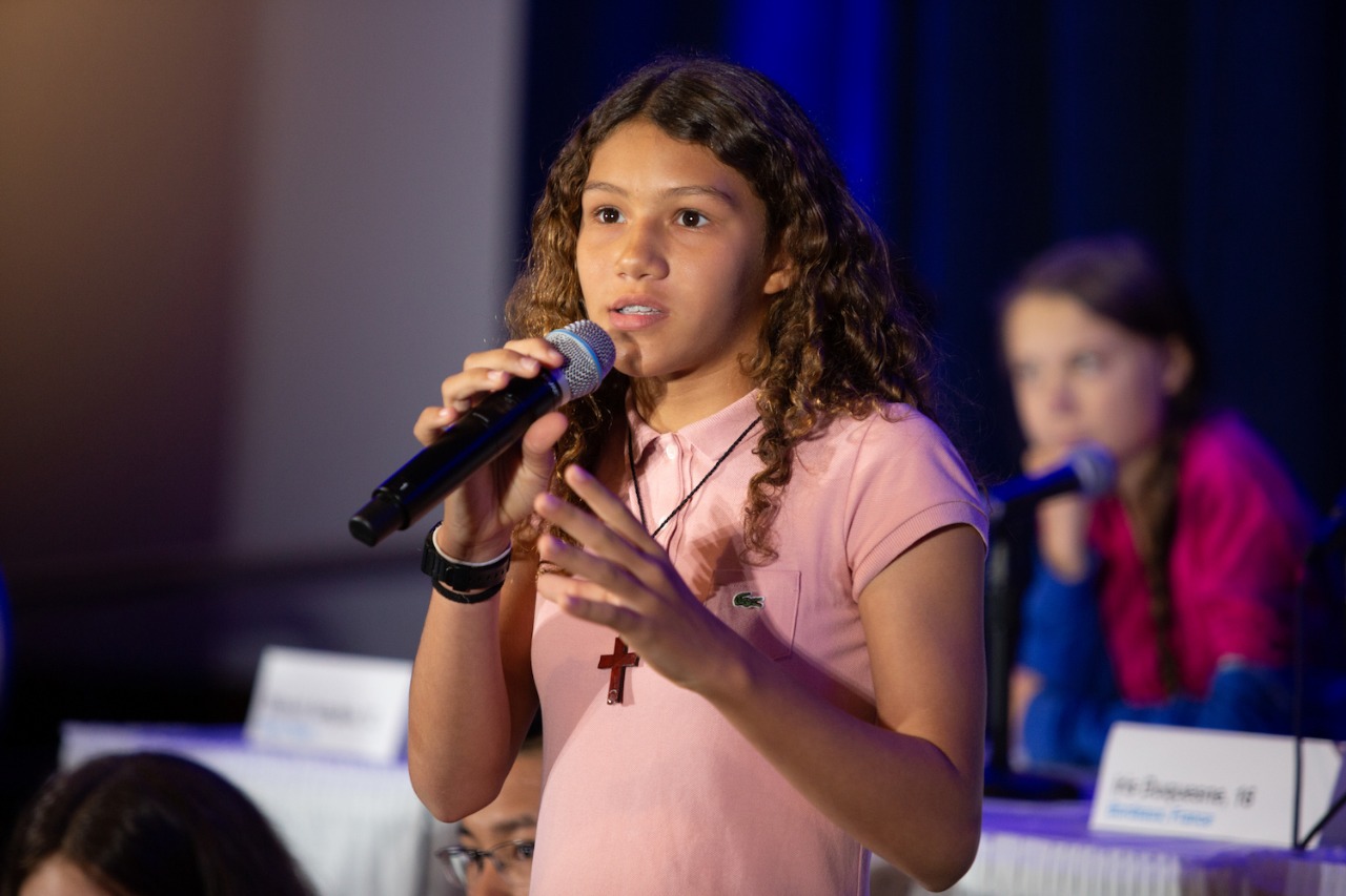 Imagem de uma menina falando ao microfone com um olhar sério. Ela esta de blusa rosa com um colar com uma cruz pendurada