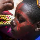 Foto de uma mulher pintando o rosto de um menino negro com tinta vermelha. Os desenhos formam um padrão tribal, com listras e círculos simétricos.