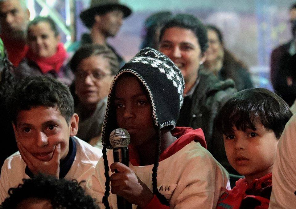 Diversas crianças sentadas assistindo a um evento literário. Em destaque, um menino negro segura um microfone.