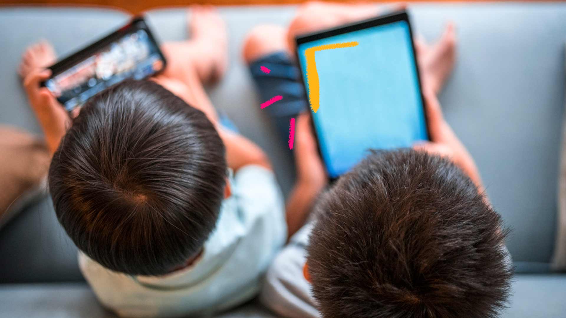 Foto captada de cima mostra dois meninos de costas sentados no sofá, cada uma com um aparelho eletrônico nas mãos. Um tablet e um celular.