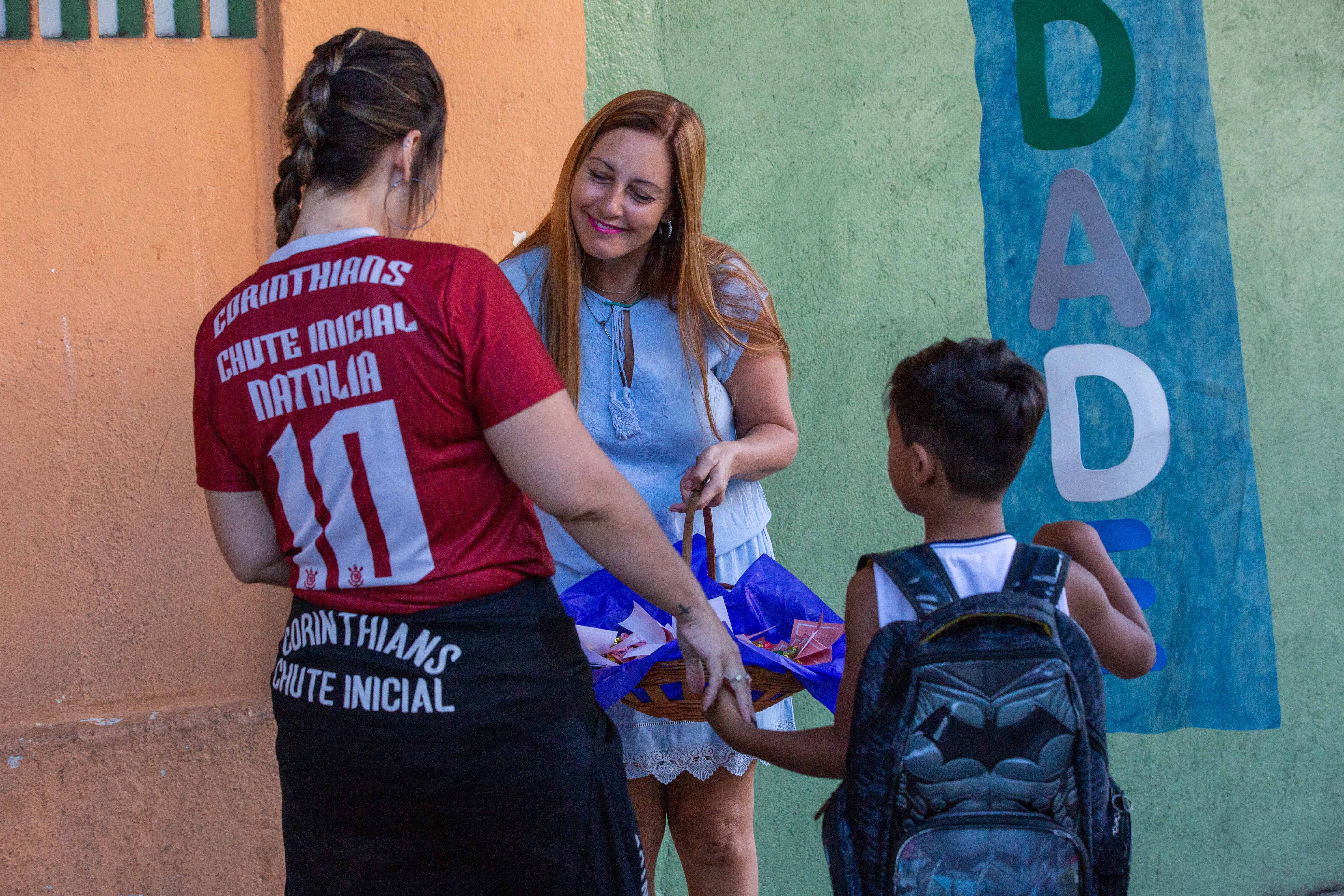 Você permite que uma professora tenha dificuldade com seu filho? Imagem de uma mãe levando seu filho para escola. A professora está sorrindo com uma cesta em suas mãos entregando um presente para a mãe