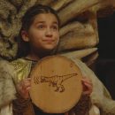 Imagem de uma menina homenageando Humboldt. Ela esta com um pedaço de madeira redondo em suas mãos com um desenho de dinossauro.