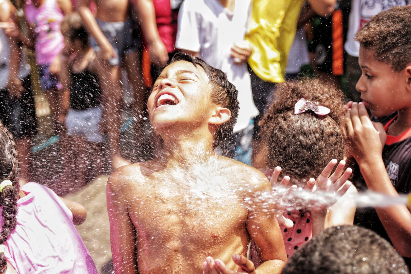 Foto de um menino sem camiseta. Estão jogando água nele, e ele está todo molhado. Logo atrás uma menina com a cabeça abaixada e as mãos em seu rosto