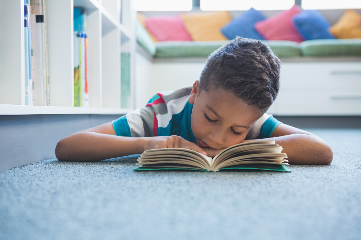 Imagem de um menino apoiado em uma mesa com a cabeça em cima dos braços lendo um livro